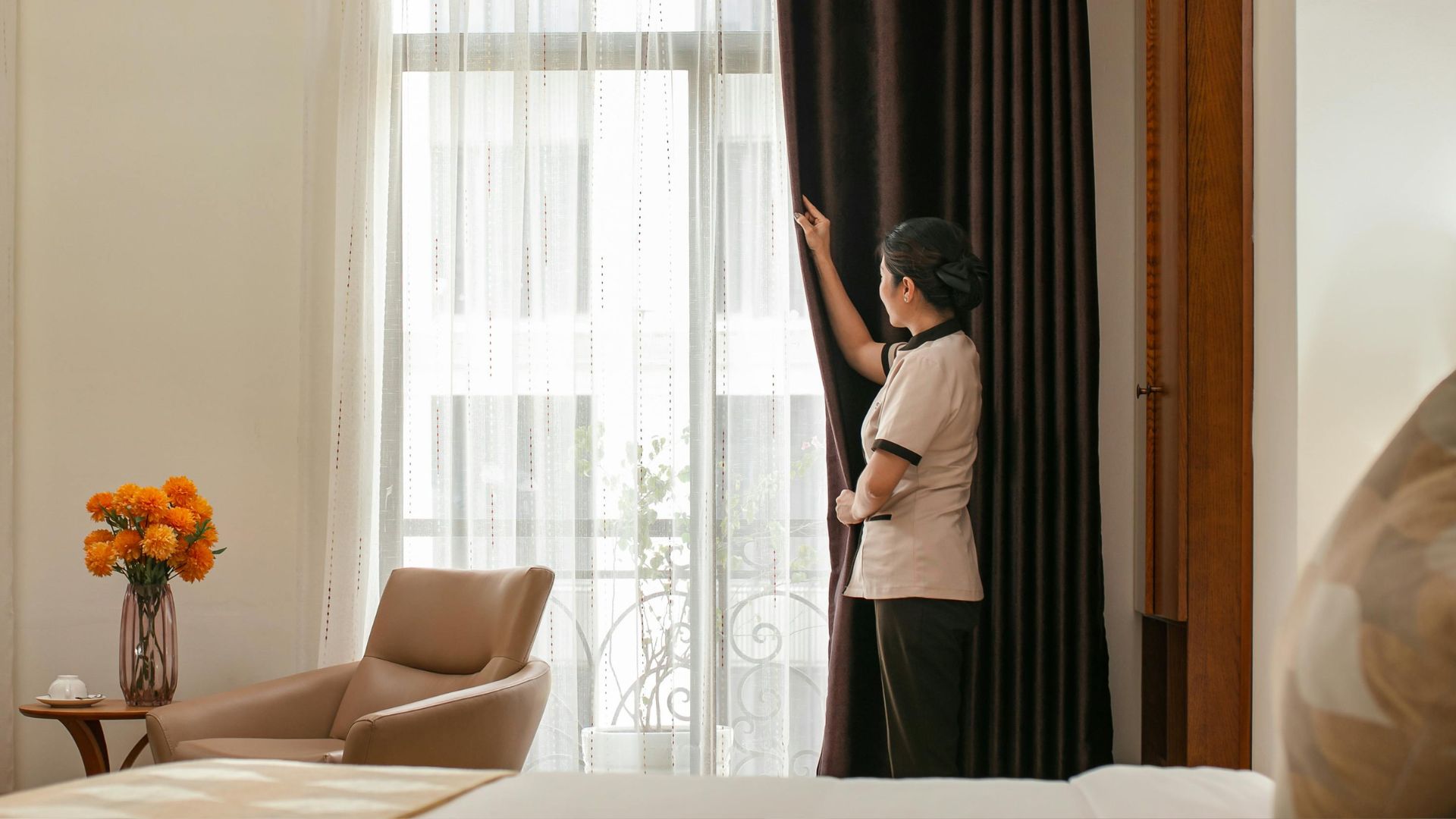 Housekeeper adjusting curtains in a bright hotel room with orange flowers