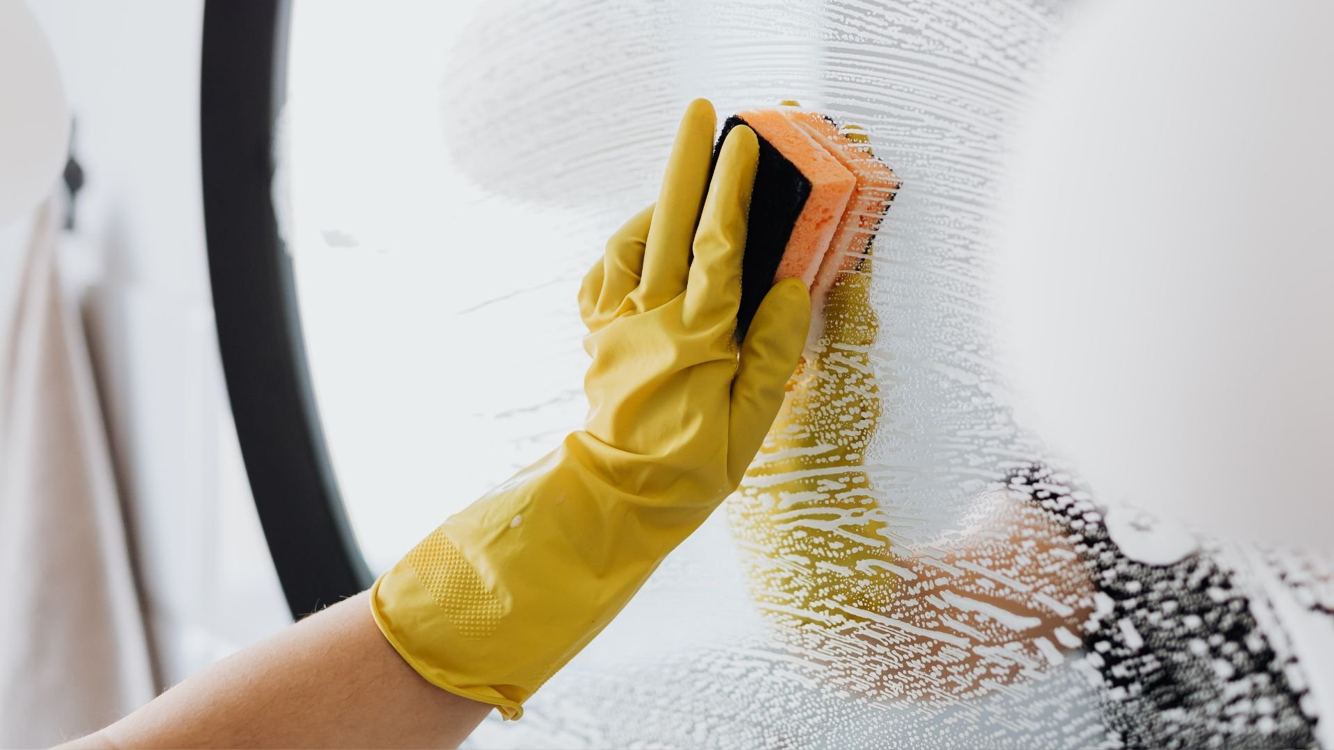 Yellow gloved hand scrubbing glass surface with orange sponge